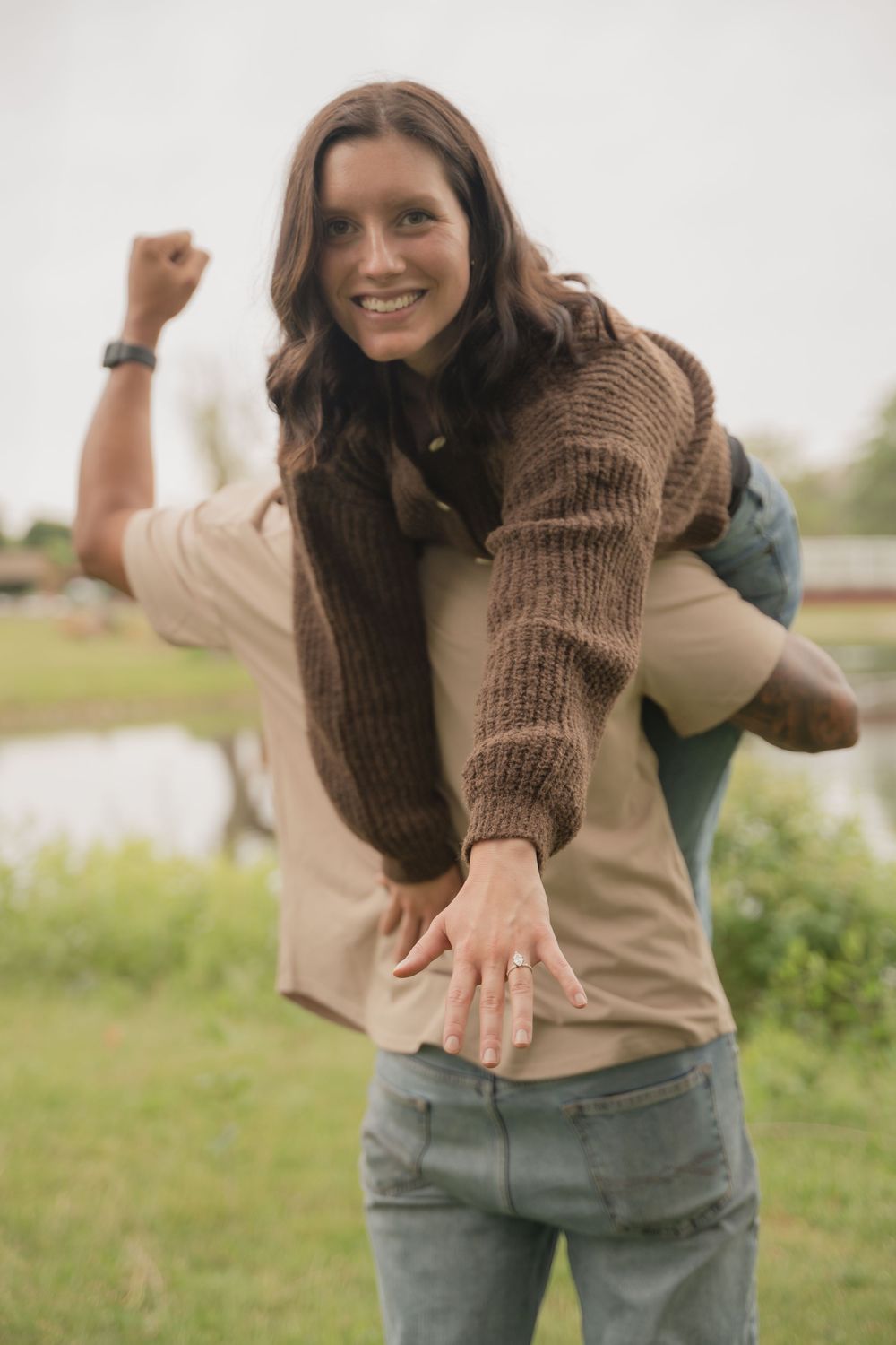 Smiling woman in brown knit sweater showing engagement ring outdoors near a lake.