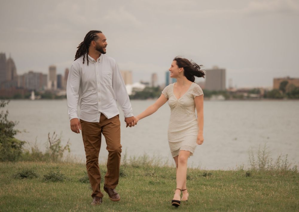 Couple holding hands walking on grass by a river with a city skyline in the background on a cloudy day.