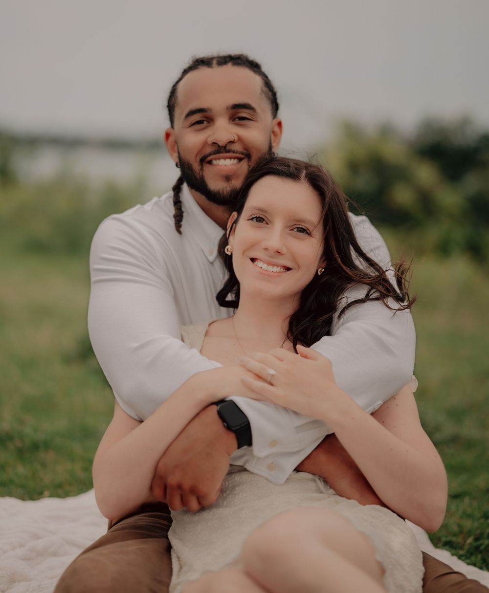 Happy couple sitting outdoors on a blanket, man embracing woman from behind, smiling near a waterfront.