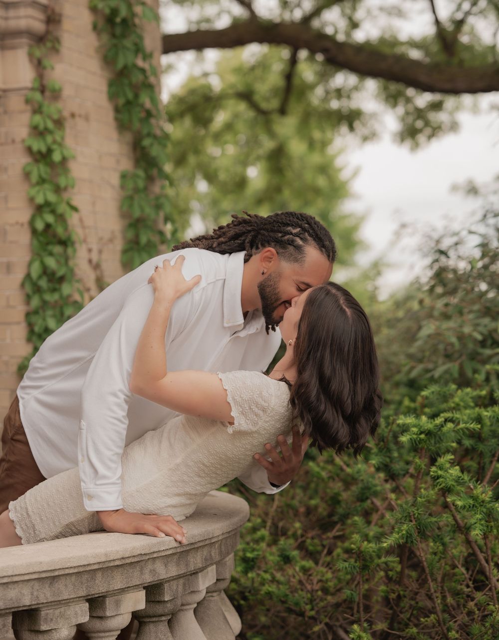 Couple sharing a romantic dip kiss on a stone balcony surrounded by lush green ivy and trees.