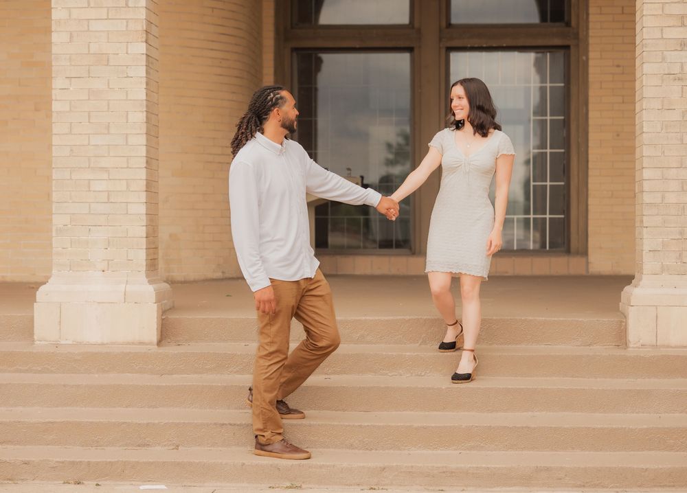 Couple holding hands on steps of a building, man in khakis and woman in white dress, outdoor engagement photo.