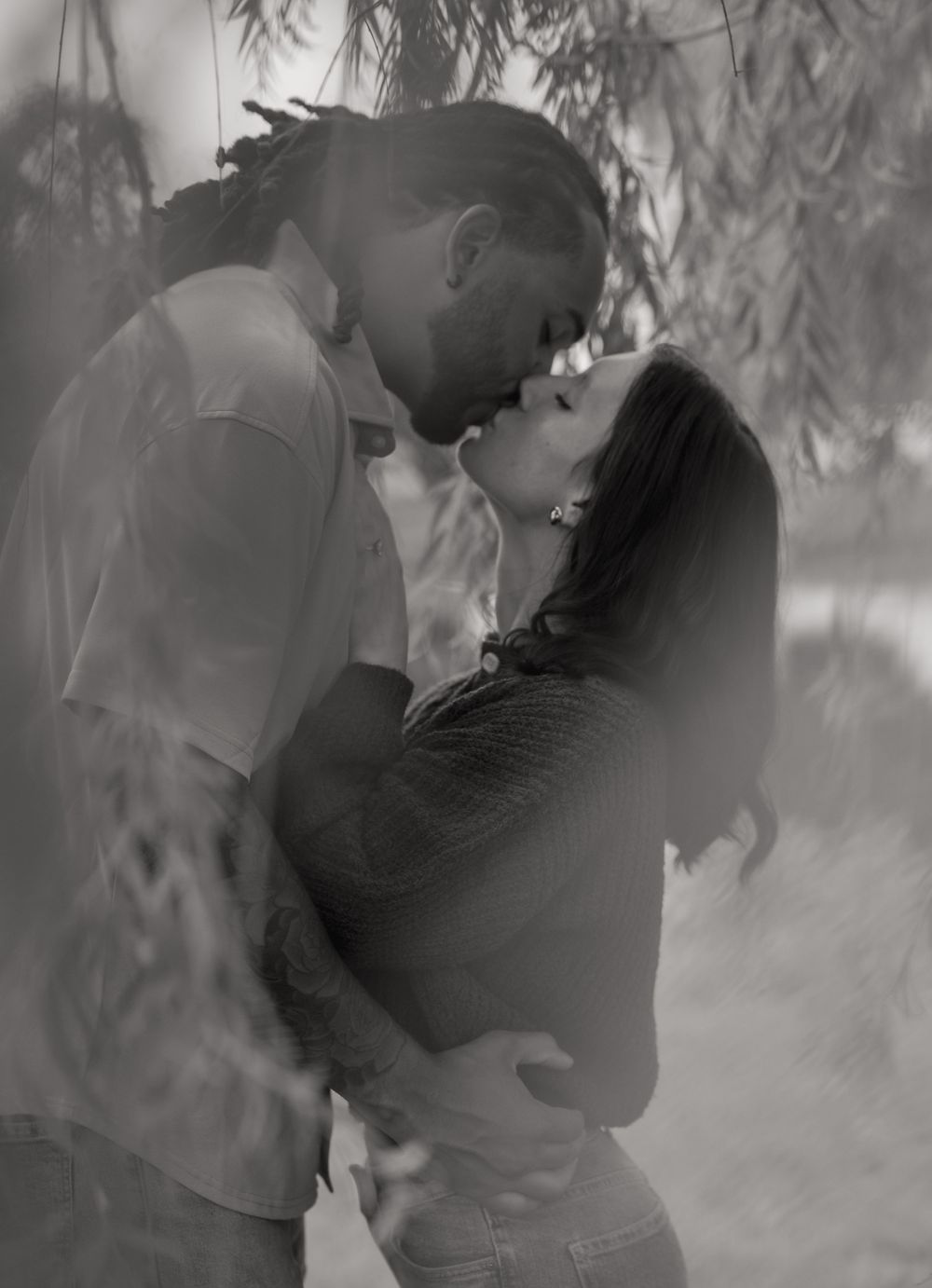 Black and white romantic photo of a couple sharing a tender kiss outdoors beneath hanging tree branches.