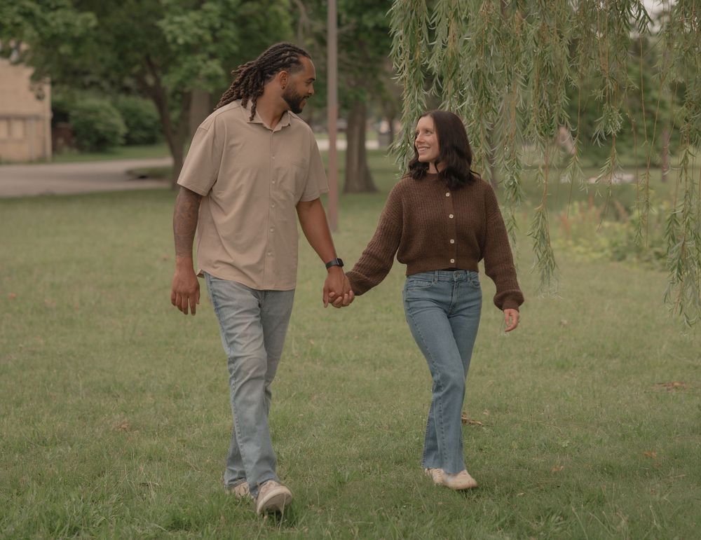 A couple holds hands and smiles while walking through a lush green park surrounded by trees.