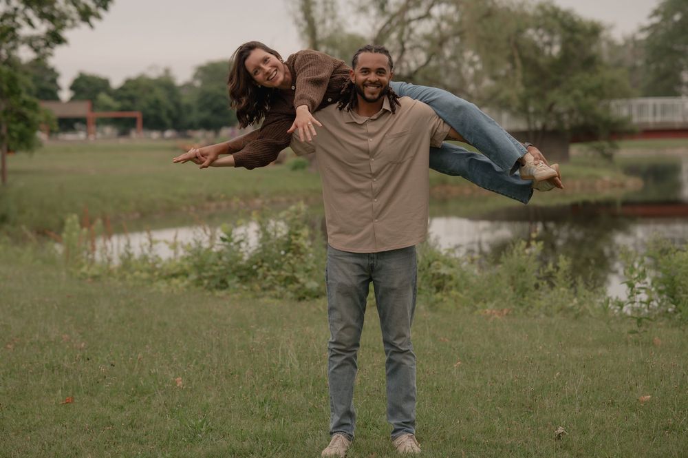 Man lifting woman over his shoulder playfully in a green park near a pond, both smiling joyfully.