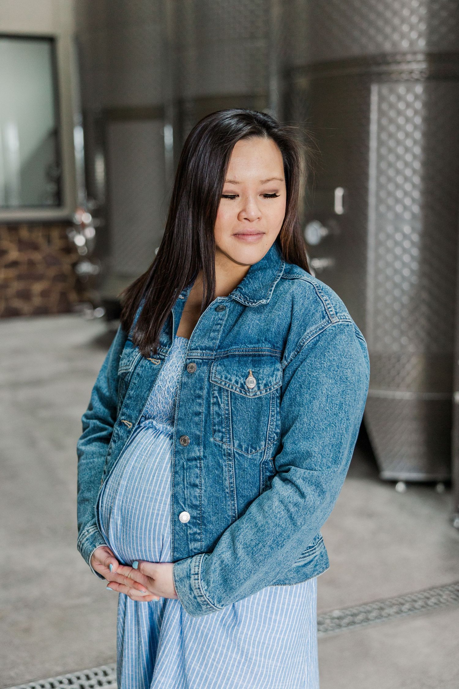 Pregnant woman in denim jacket and blue dress cradling baby bump, standing in modern winery with steel fermentation tanks.