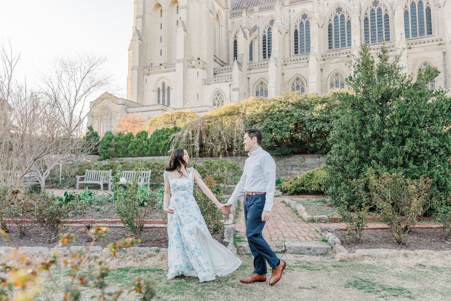 Couple holds hands walking in garden before grand cathedral during outdoor engagement photo session.
