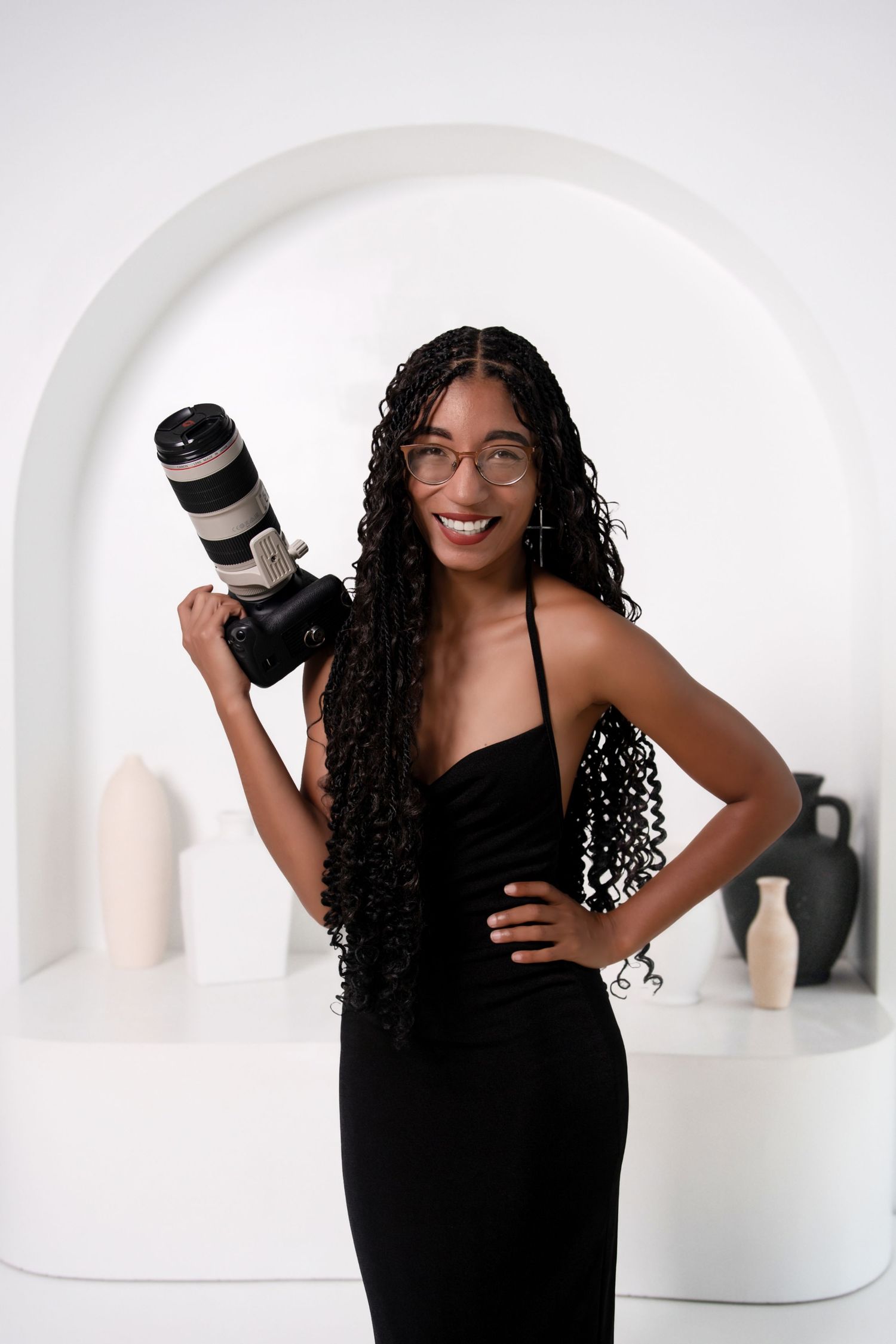 Smiling Black woman photographer in black dress and glasses holding a Canon camera with telephoto lens in a white studio.