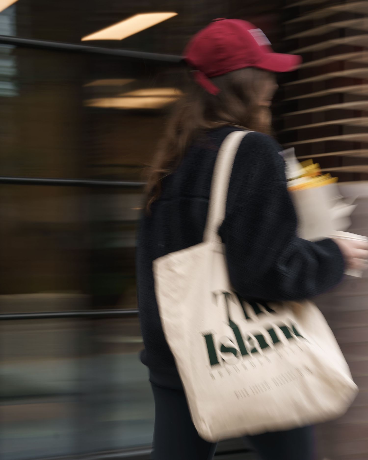 Woman in red cap and black jacket carrying a cream tote bag with text while walking in a blurred motion.
