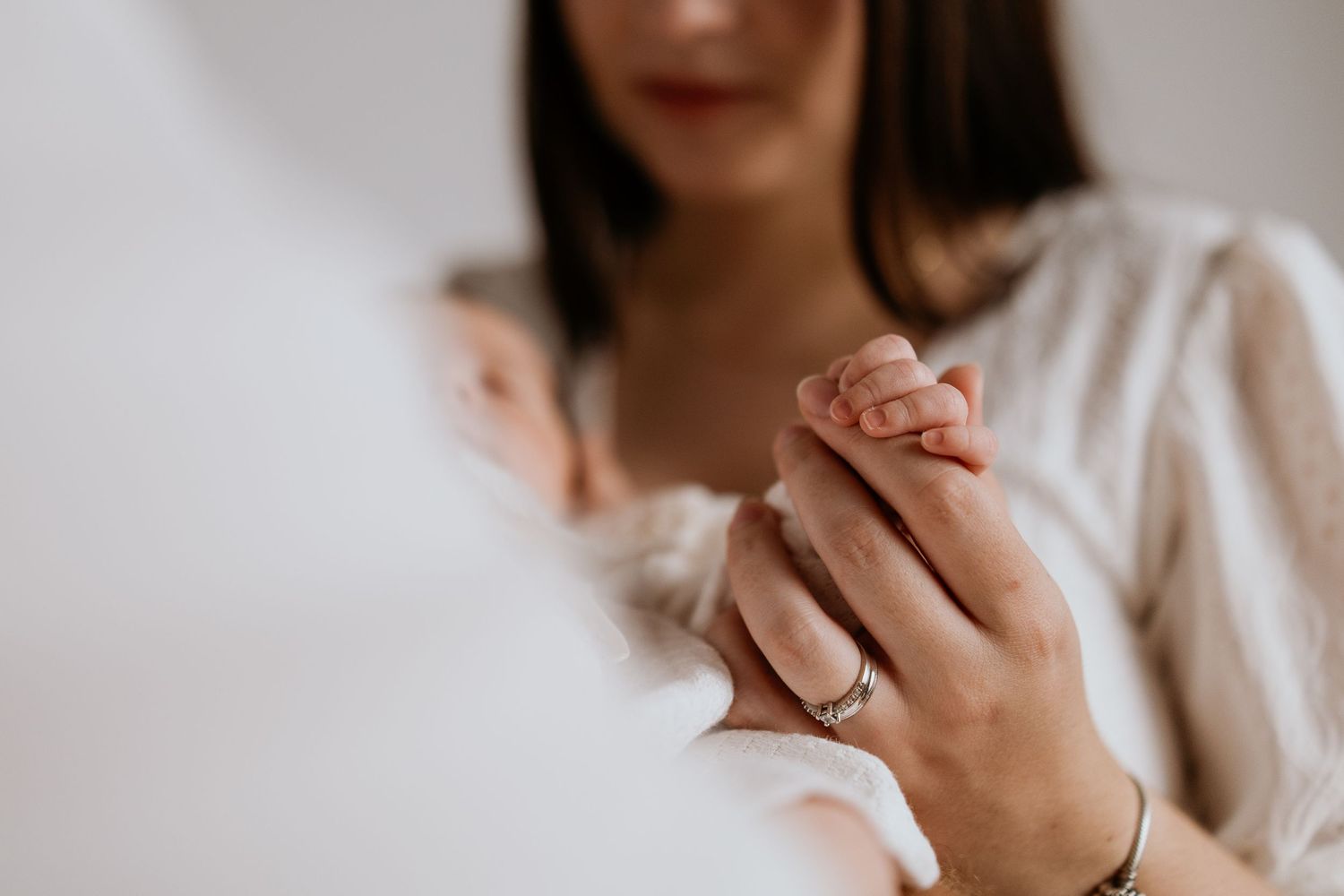 A mother gently holds her newborn baby's tiny hand, wearing a silver ring and bracelet, in a soft, tender moment.