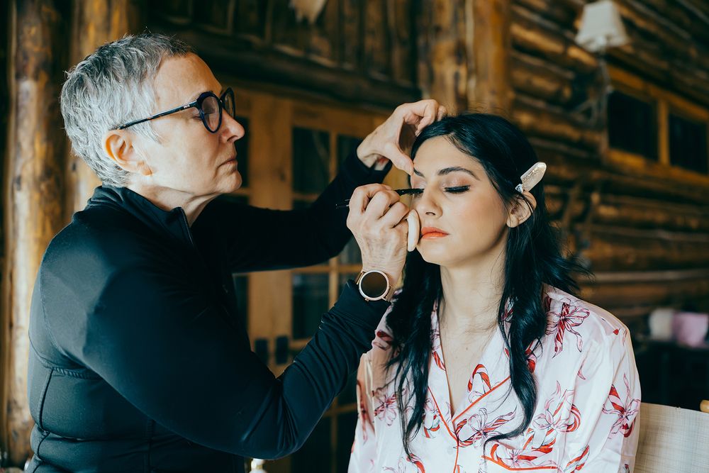 A makeup artist applies eyeshadow to a dark-haired woman in a floral robe inside a rustic log cabin.
