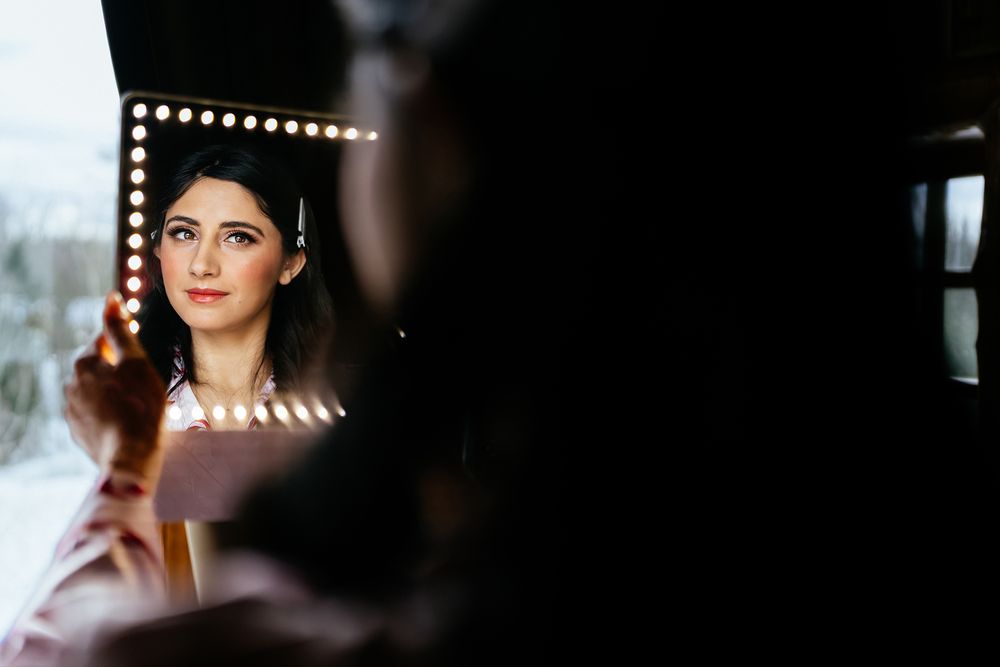 Bride getting makeup done, reflected in a lit vanity mirror with warm bokeh lights framing her face.