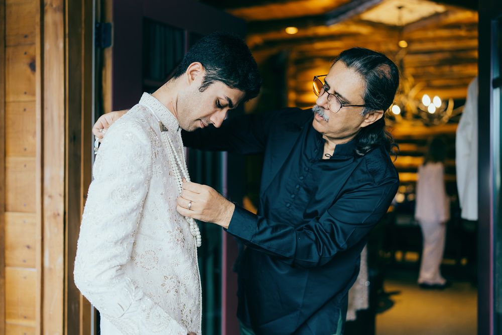 Man in black outfit helping groom dress in elegant white sherwani inside a wooden venue.