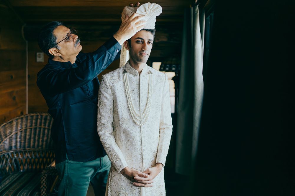 A man in traditional white Indian sherwani gets his turban placed by another man in a rustic wooden interior.