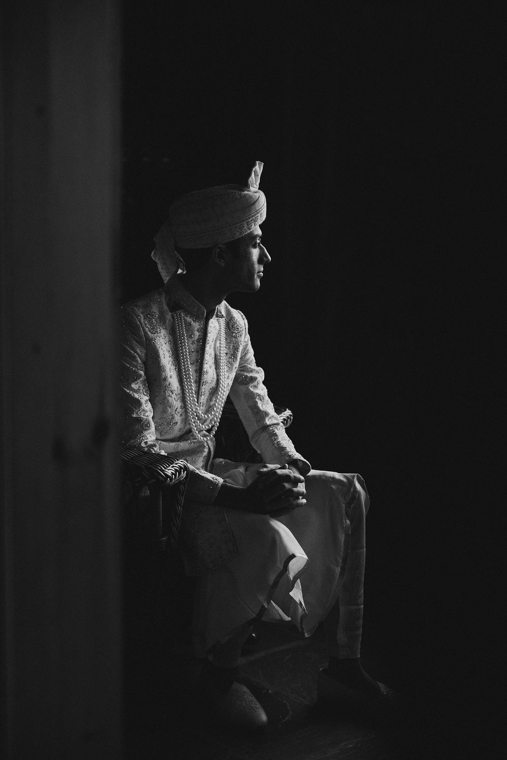 Black and white portrait of an Indian groom in traditional attire, turban, and garlands sitting contemplatively.