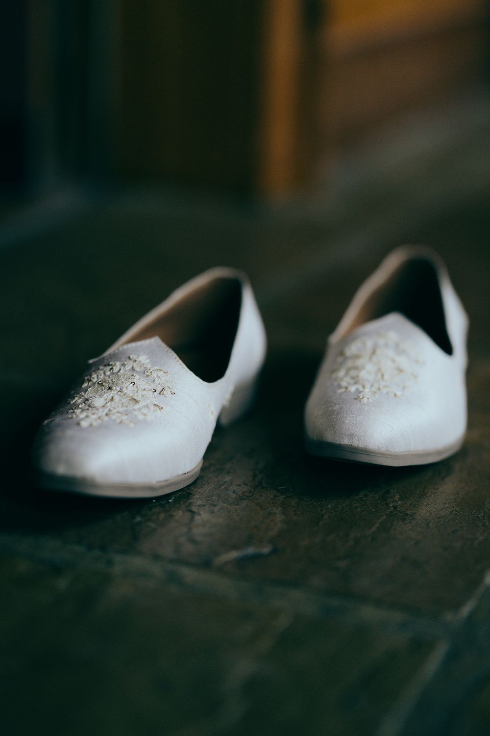 White satin bridal flats with pearl and crystal embellishments displayed on dark wooden floor.