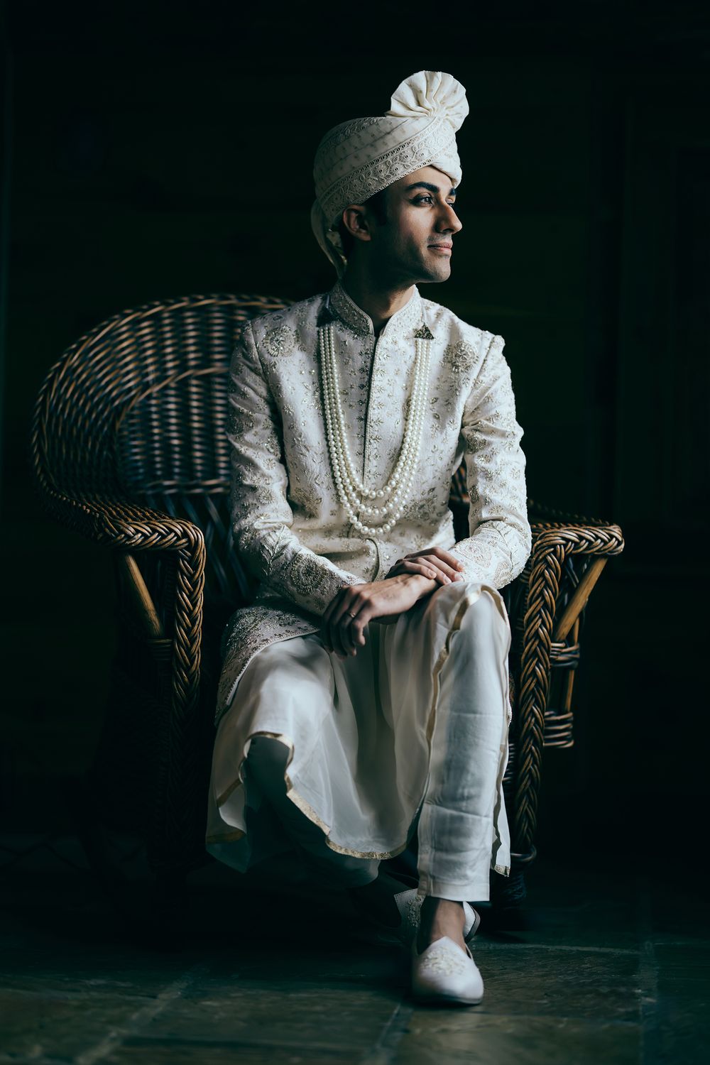 Indian groom in white sherwani and turban sits elegantly in wicker chair against dark background.