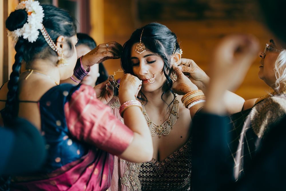 Indian bride being adorned with traditional jewelry by bridesmaids in colorful saris during wedding preparation.