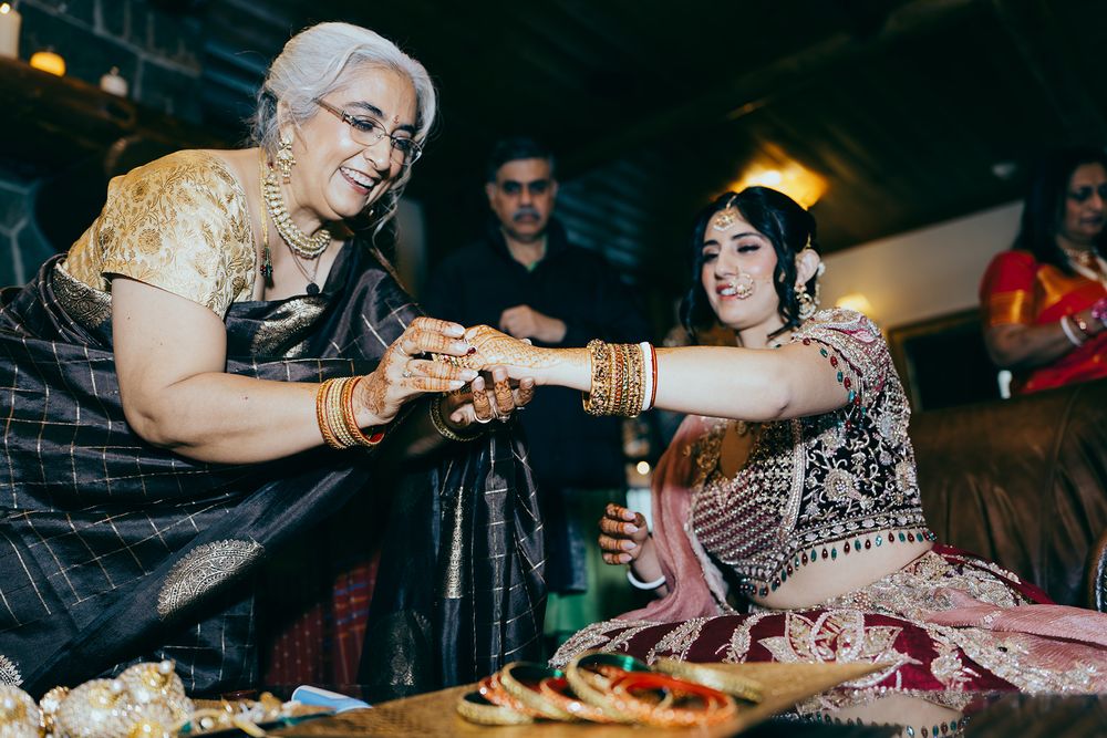 Elderly woman in black saree exchanges jewelry with bride in pink lehenga during Indian wedding ceremony.