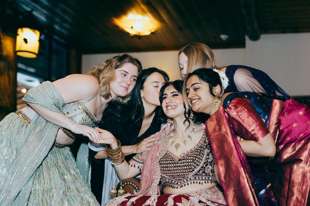 Bride in ornate lehenga surrounded by bridesmaids giving kisses at Indian wedding celebration.