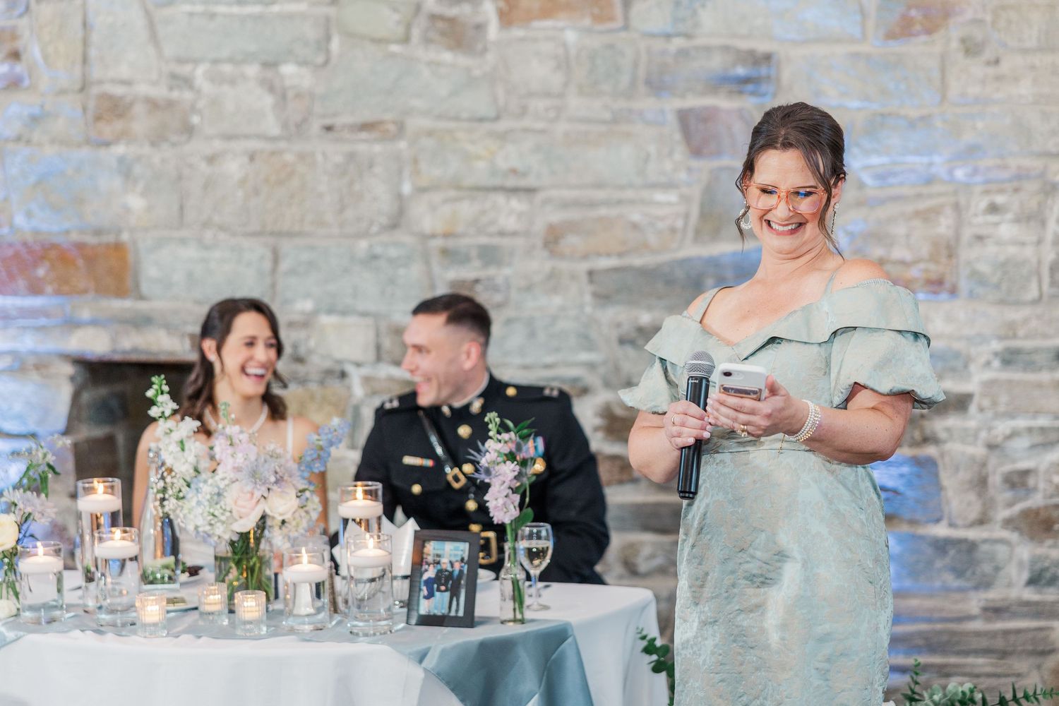 Woman in blue off-shoulder gown gives wedding toast while bride and groom in military uniform smile at reception table.