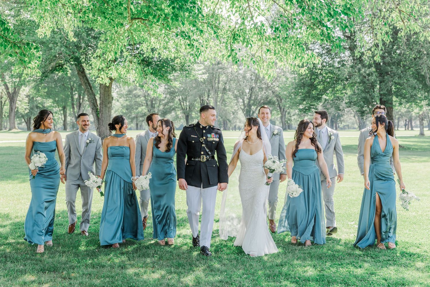 Wedding party walks outdoors under green trees, bridesmaids in blue dresses, groomsmen in gray suits, groom in military uniform.
