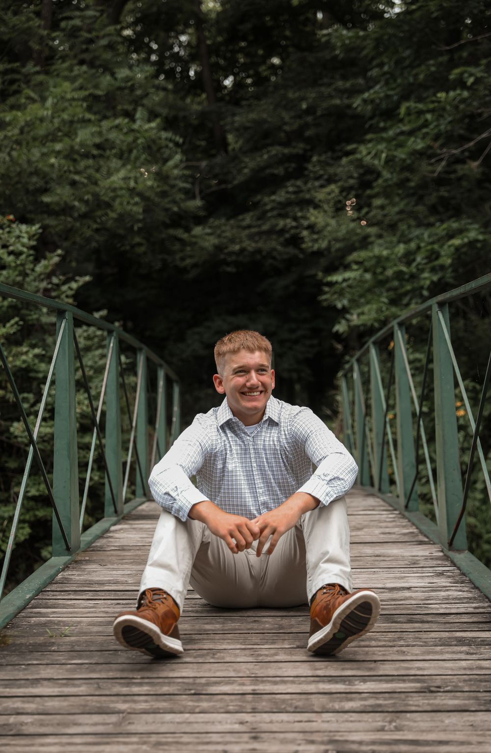 Young man smiling on a wooden bridge surrounded by lush green trees, wearing a light blue shirt and white pants.