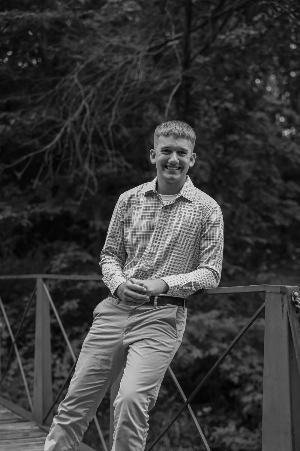 Young man smiling on a wooden bridge surrounded by trees, wearing a button-up shirt and chinos, black and white photo.