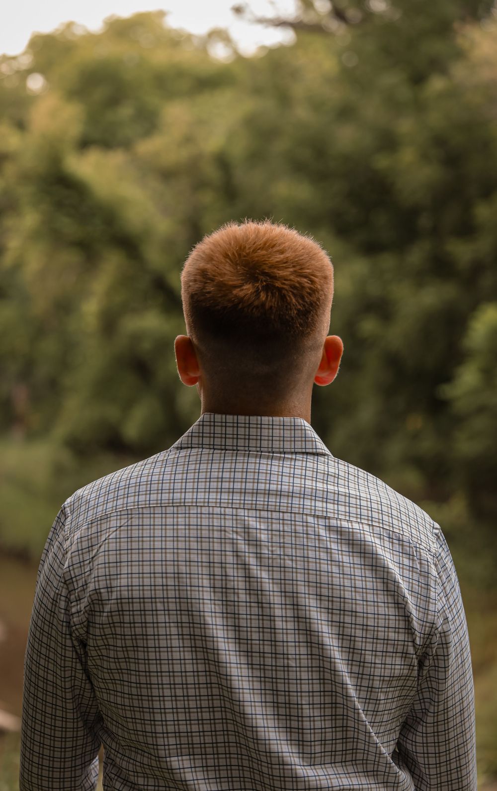 Young man with red hair wearing a plaid shirt, viewed from behind, standing outdoors near green trees.