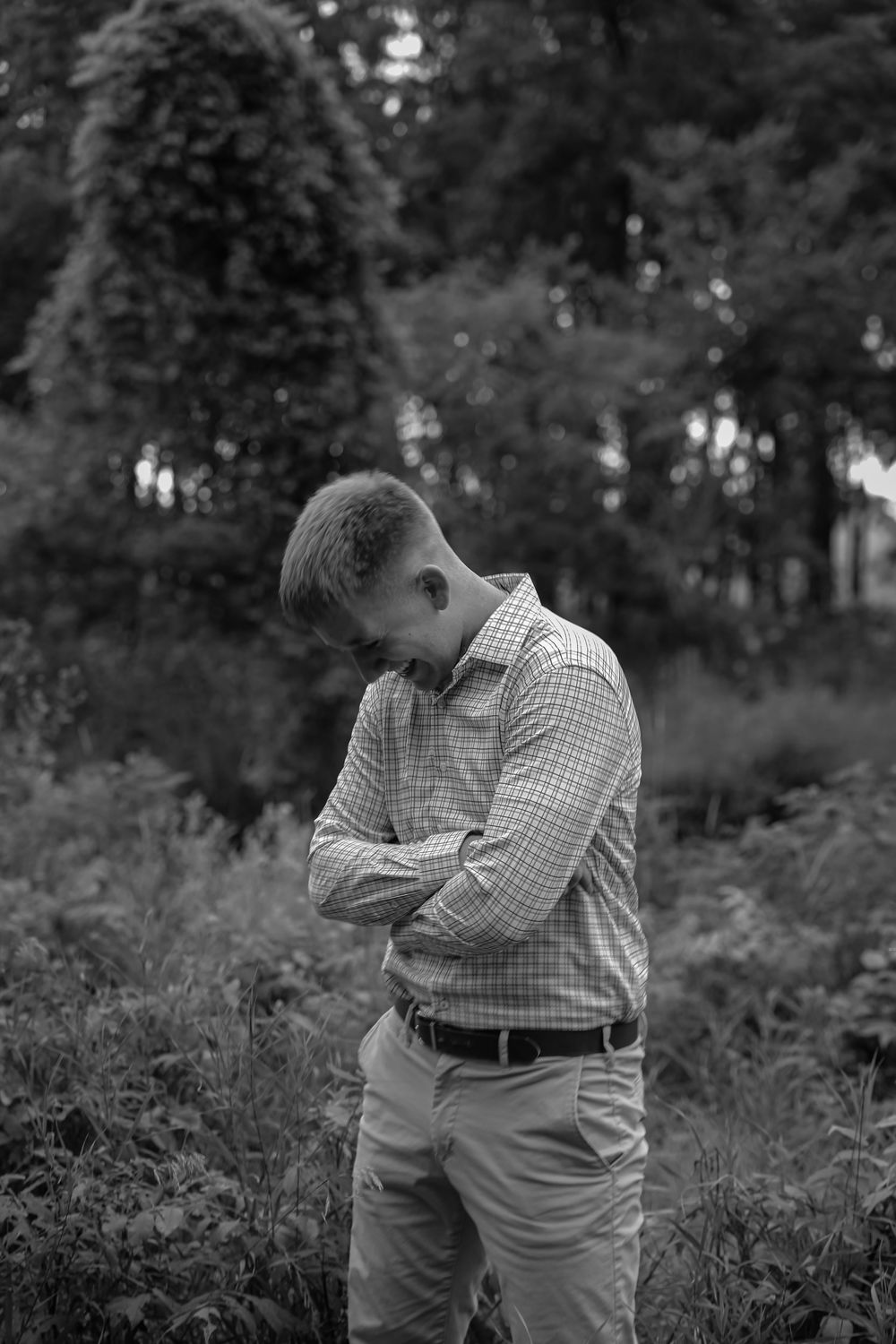 Young man in plaid shirt stands with arms crossed looking down in a black and white outdoor portrait.
