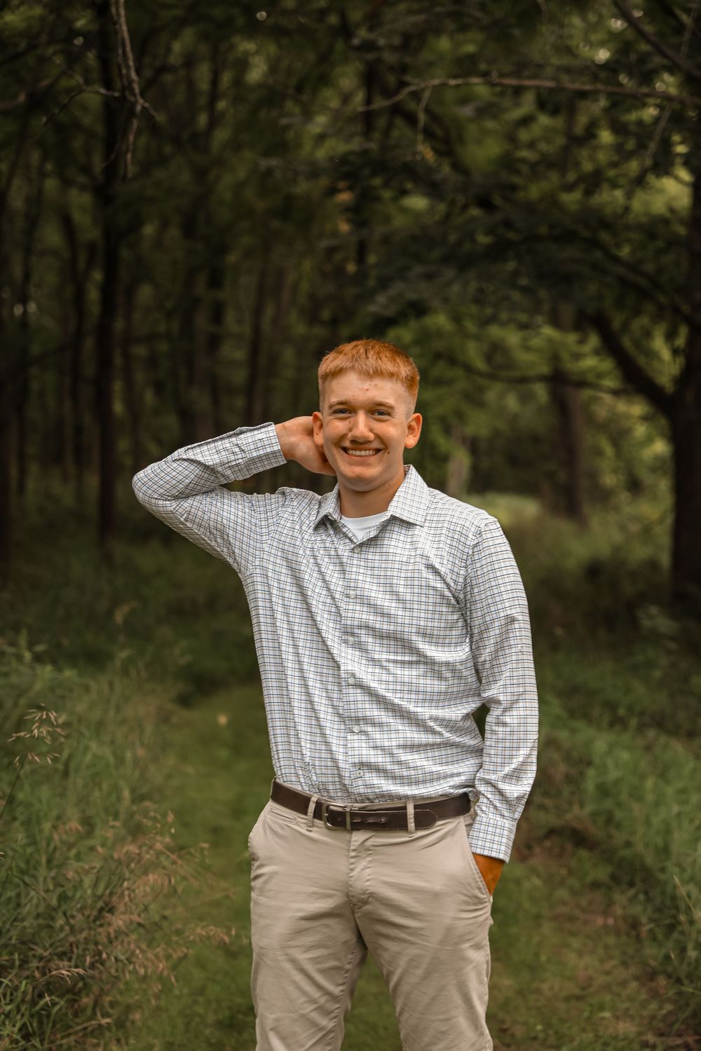 Smiling young man with red hair in a plaid shirt and khakis posing on a forest path.