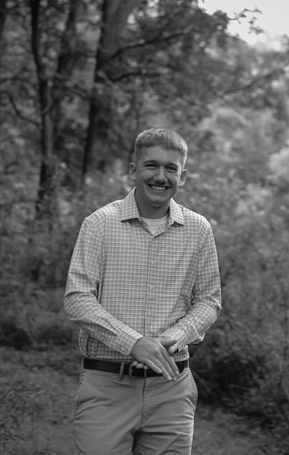 Smiling young man in plaid shirt standing outdoors in a wooded area, black and white portrait photo.