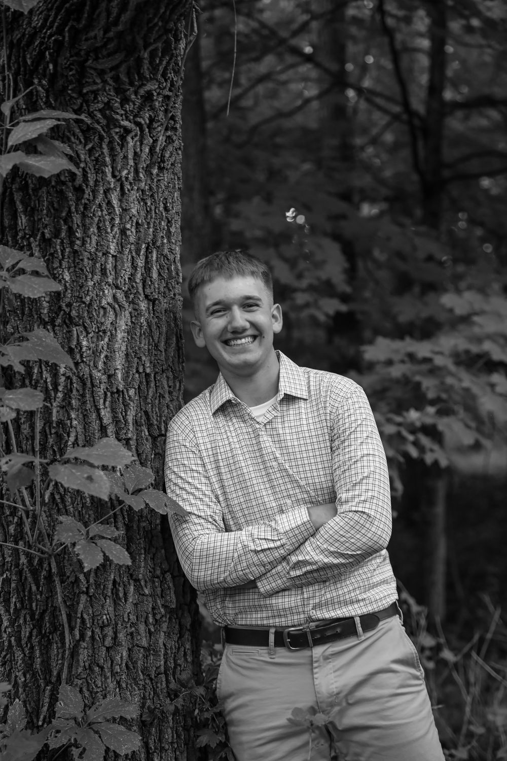 Young man in plaid shirt smiling with arms crossed leaning against a tree in a forest, black and white photo.