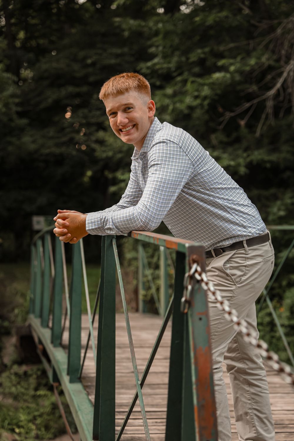 Smiling young man in plaid shirt leans on green bridge railing surrounded by lush forest.