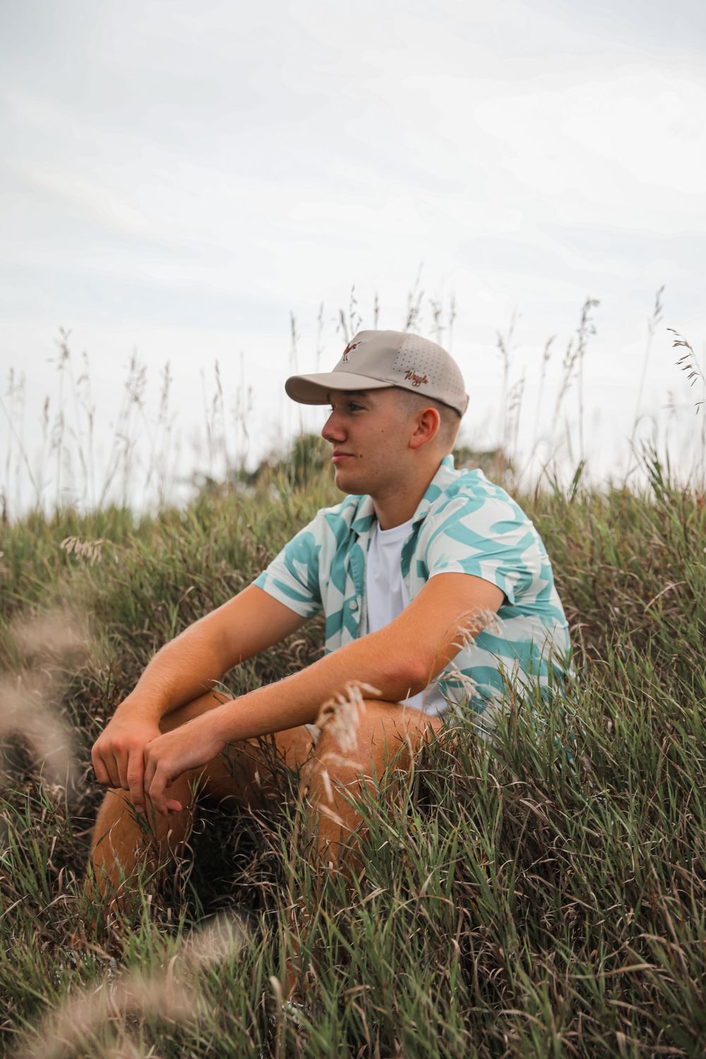 Young man in a blue patterned shirt and cap sitting in tall green grass field under a cloudy sky.