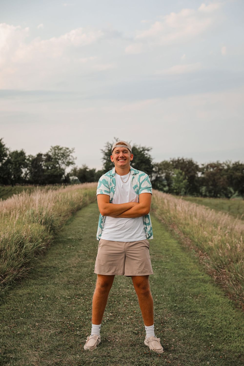 Smiling young man standing with arms crossed on a rural path surrounded by tall grass and trees under cloudy sky.
