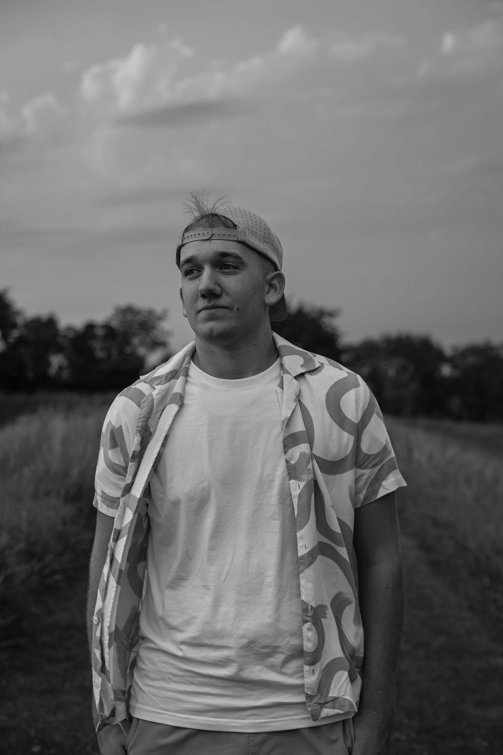 Young man in patterned shirt and backward cap stands in a field under cloudy sky in black and white photo.