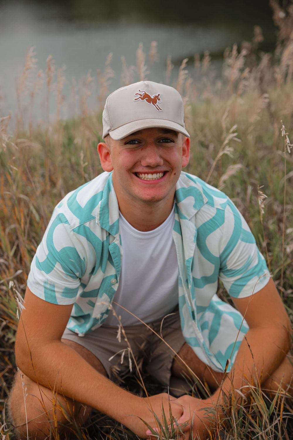 Smiling young man in a teal patterned shirt and cap sits in tall grass near a lake.