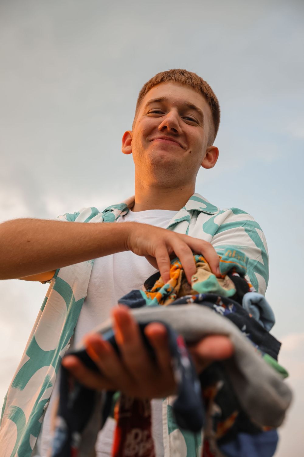 Young man smiling holding a bundle of colorful items toward camera, shot from low angle against cloudy sky.