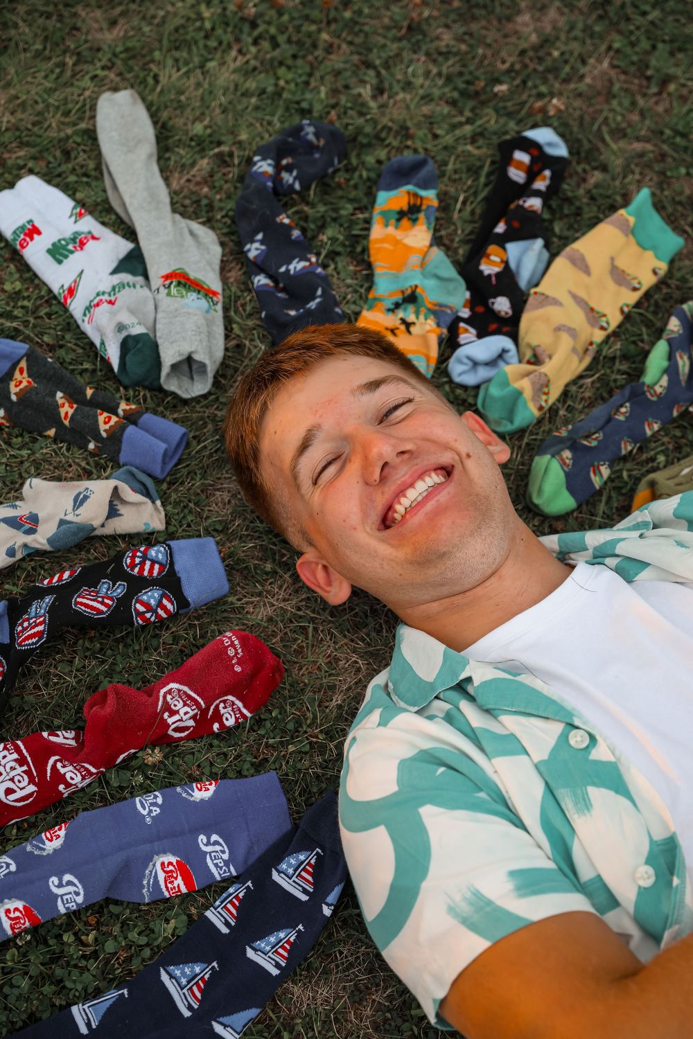 Laughing young man lies on grass surrounded by colorful patterned socks arranged around his head.