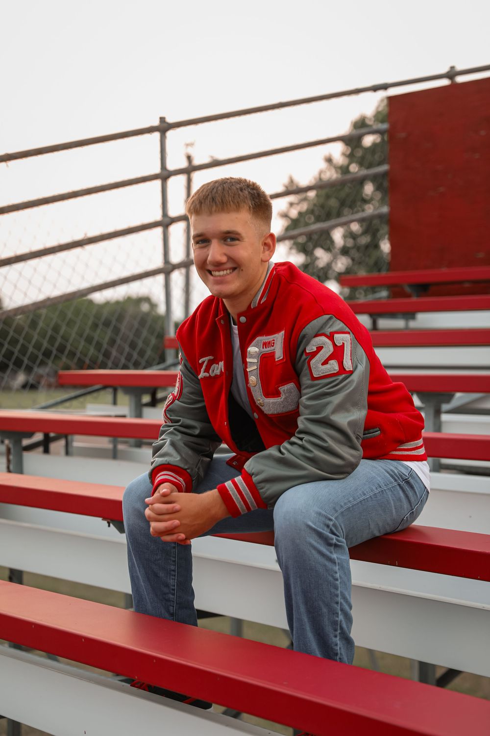 Smiling teen in red varsity letterman jacket sits on bleachers for senior portrait photo.