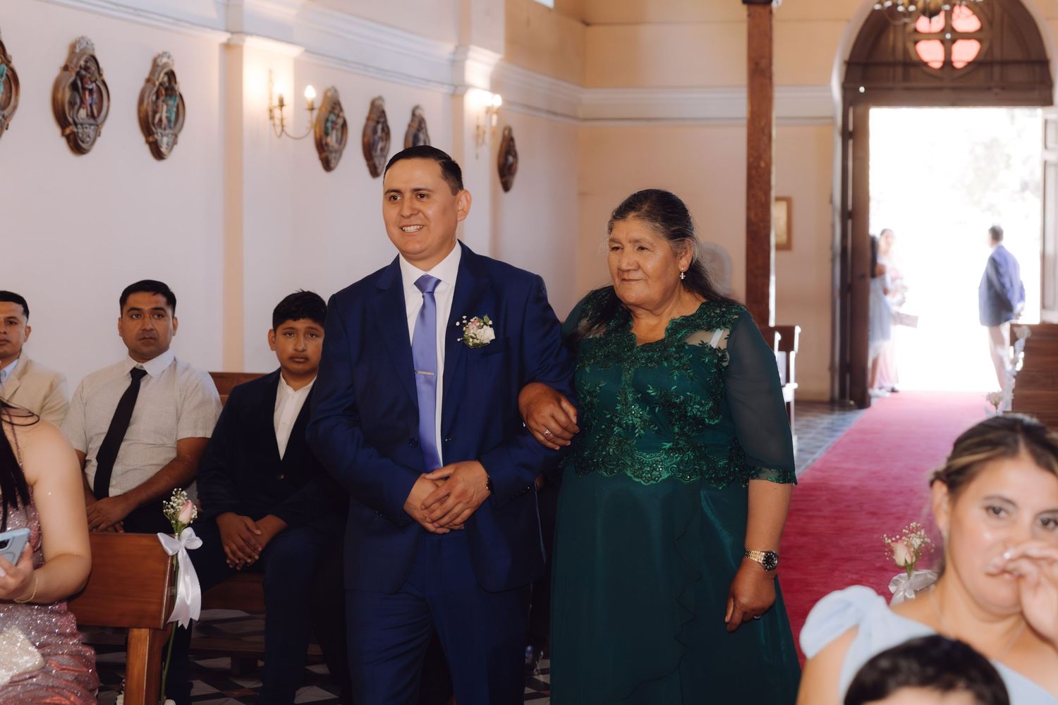 Elegantly dressed couple walking down church aisle during wedding ceremony with seated guests.