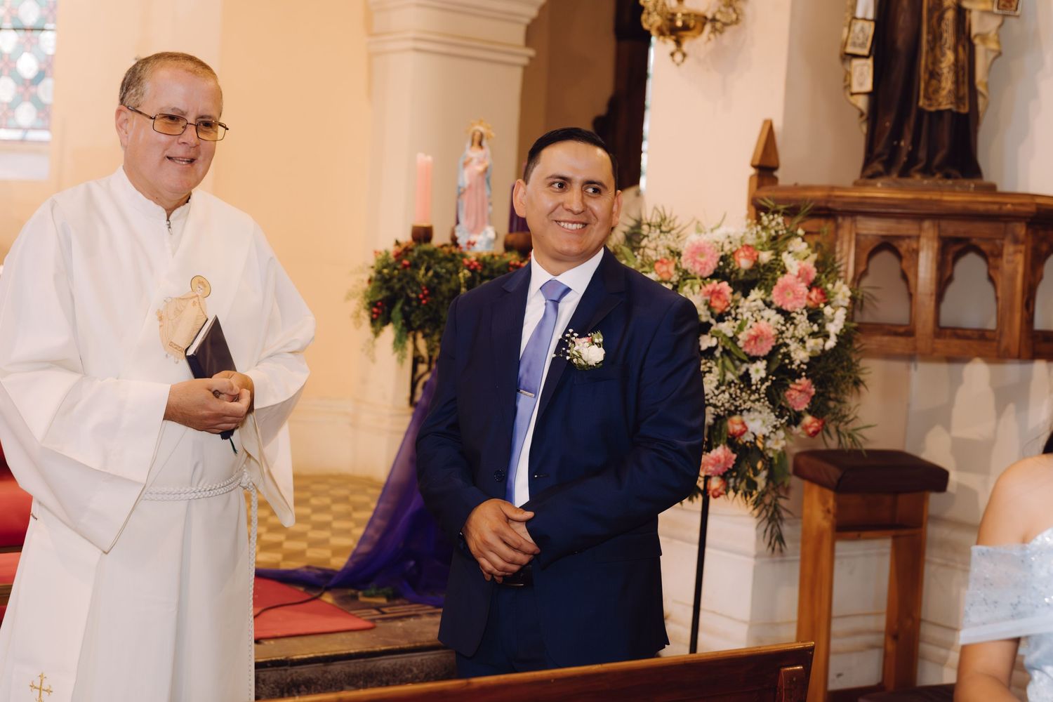 Groom in blue suit standing at church altar with priest during wedding ceremony.