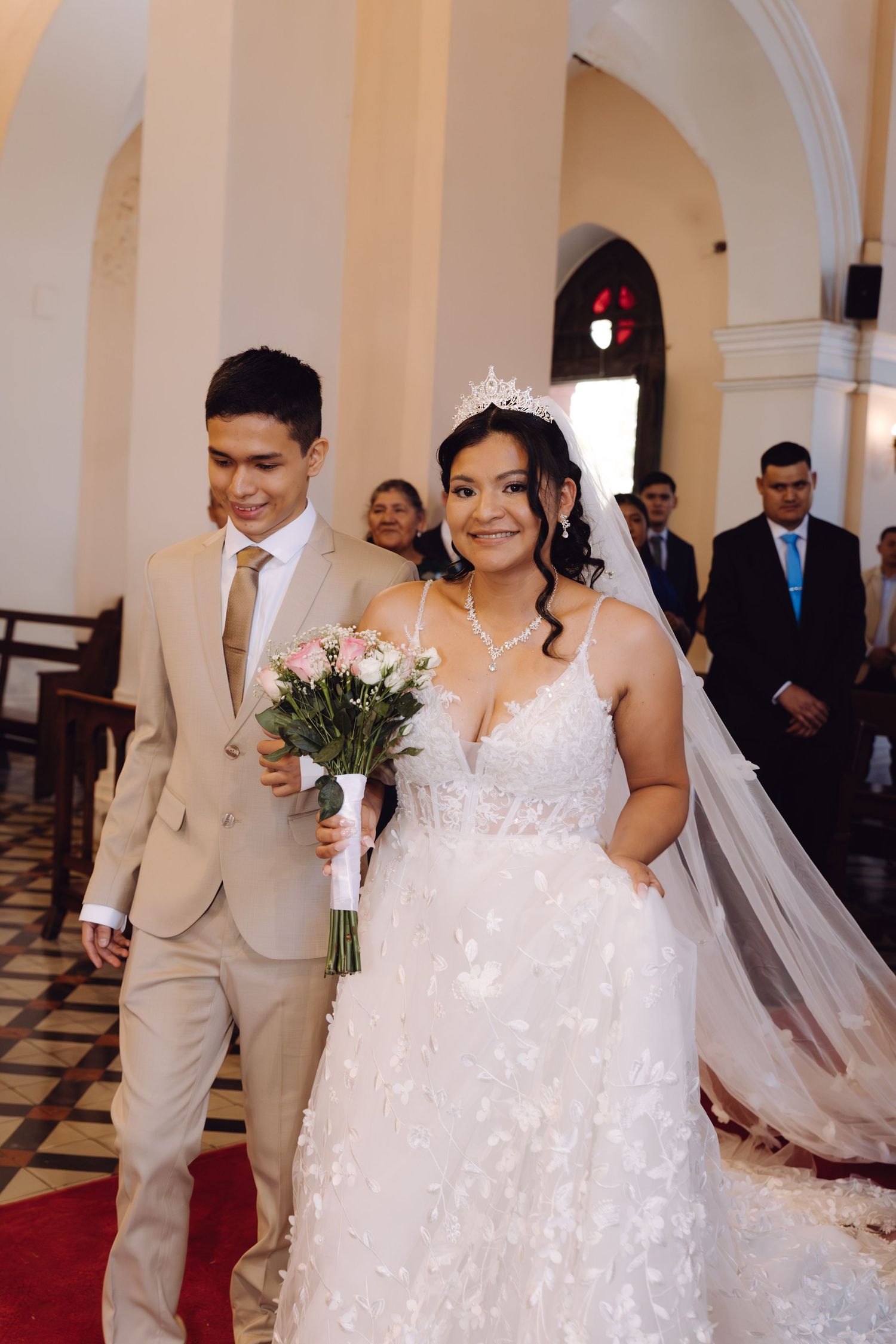 Smiling bride in tiered gown with tiara holding bouquet walking down church aisle.