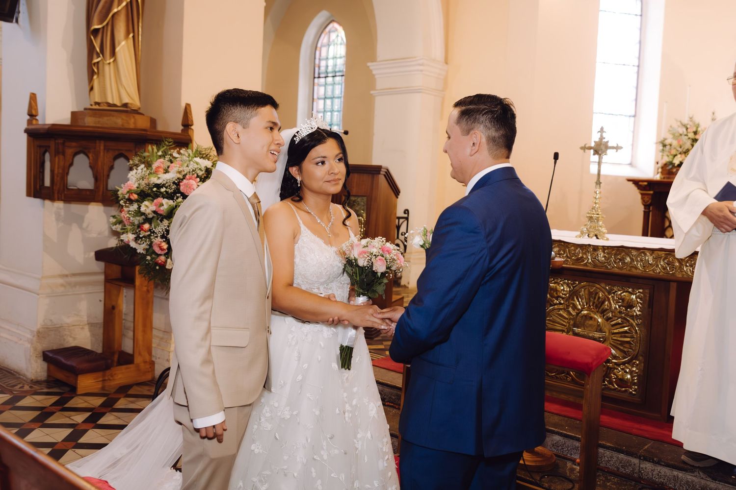 Bride in white gown holds pink bouquet while groom in tan suit and man in blue suit stand at church altar.