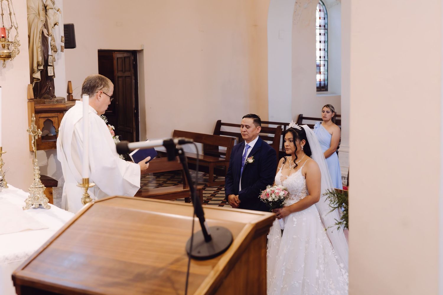 Bride in white lace gown walks down church aisle with escort toward priest standing at the altar.