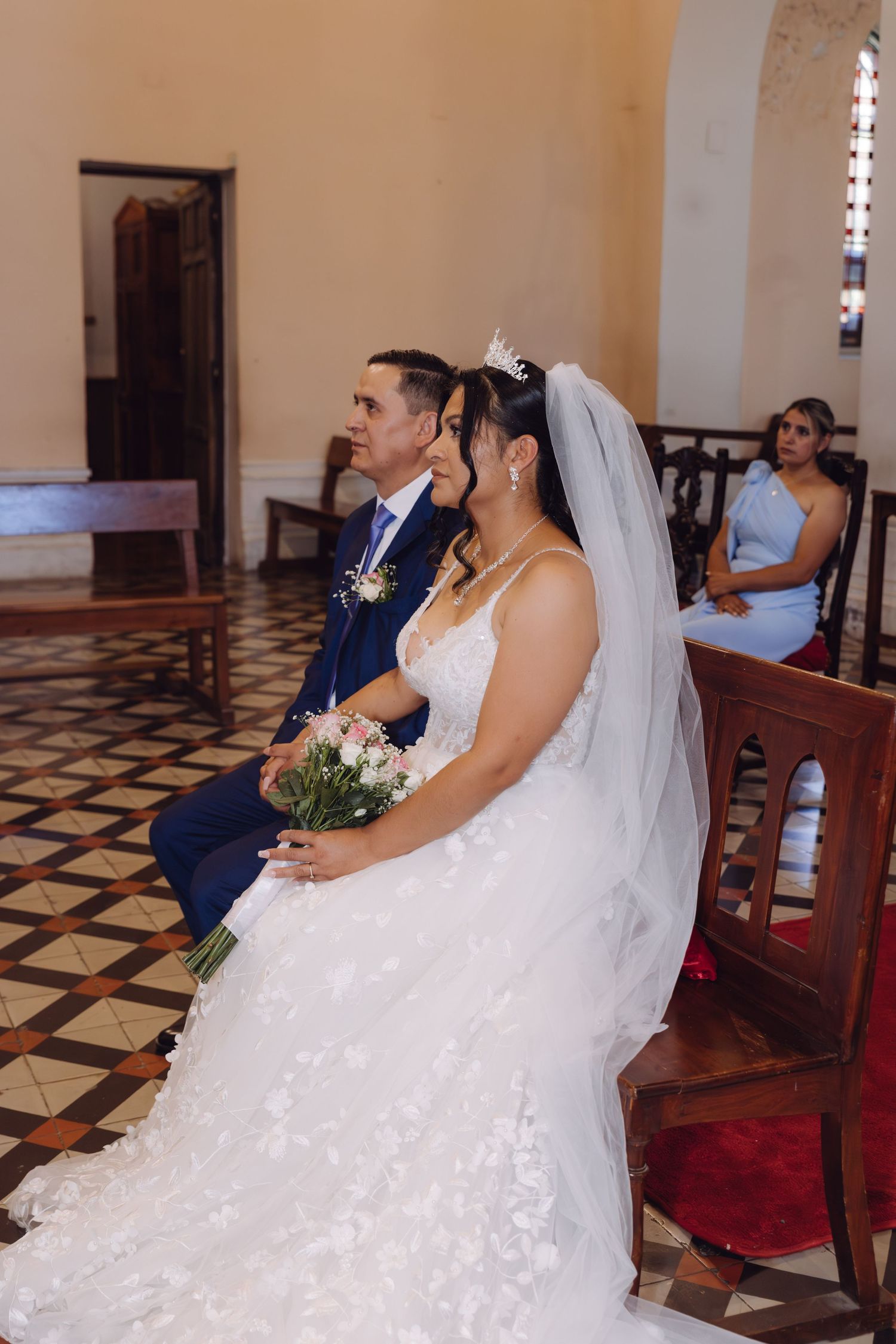 Smiling bride in white veil holds pink bouquet while seated beside groom in blue suit during church ceremony.