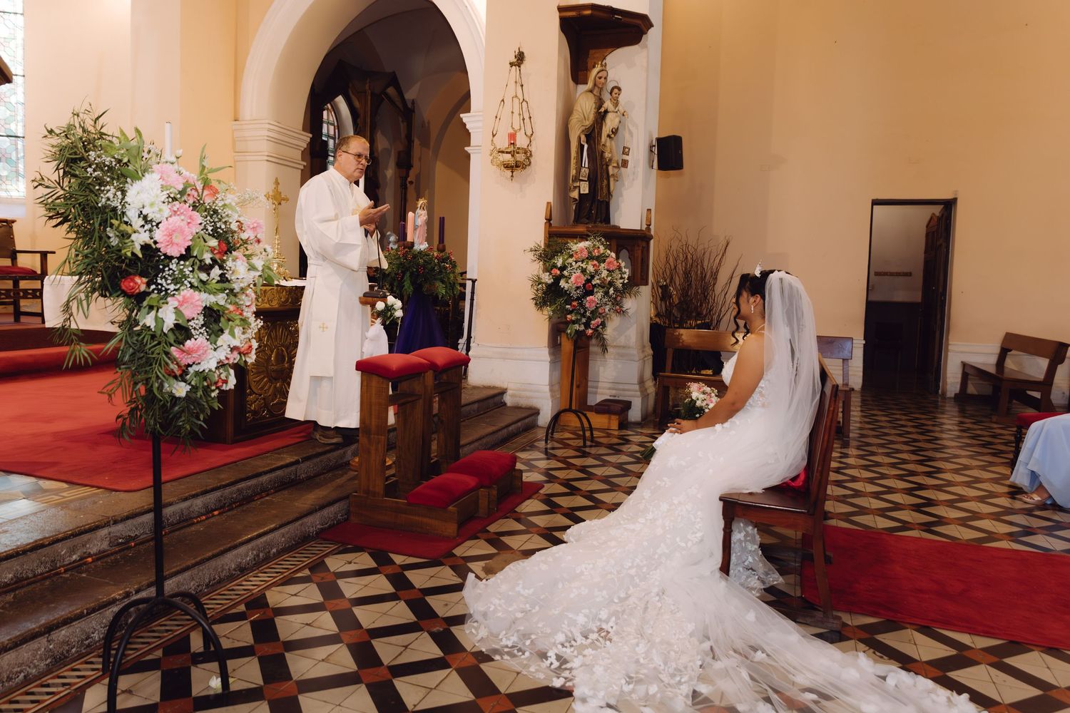 Bride in white lace wedding gown kneels before priest reading from book during Catholic church ceremony.