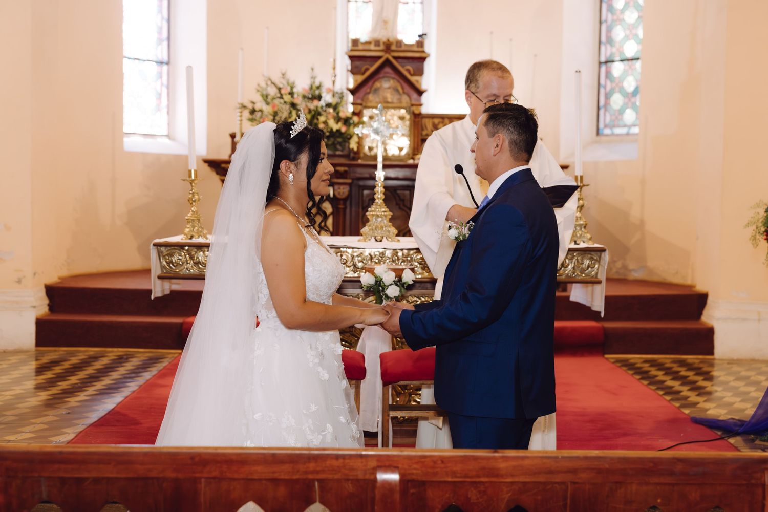 Bride and groom hold hands at church altar while priest officiates their wedding ceremony.