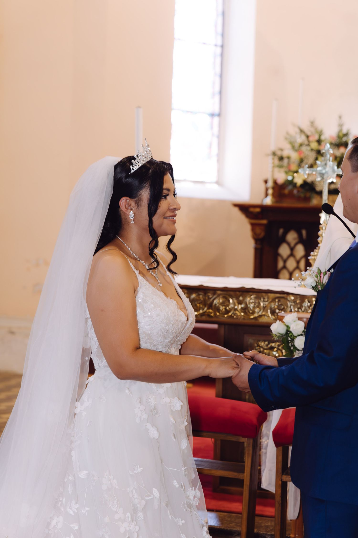 Smiling bride wearing tiara and white lace gown holds grooms hands during church wedding ceremony.
