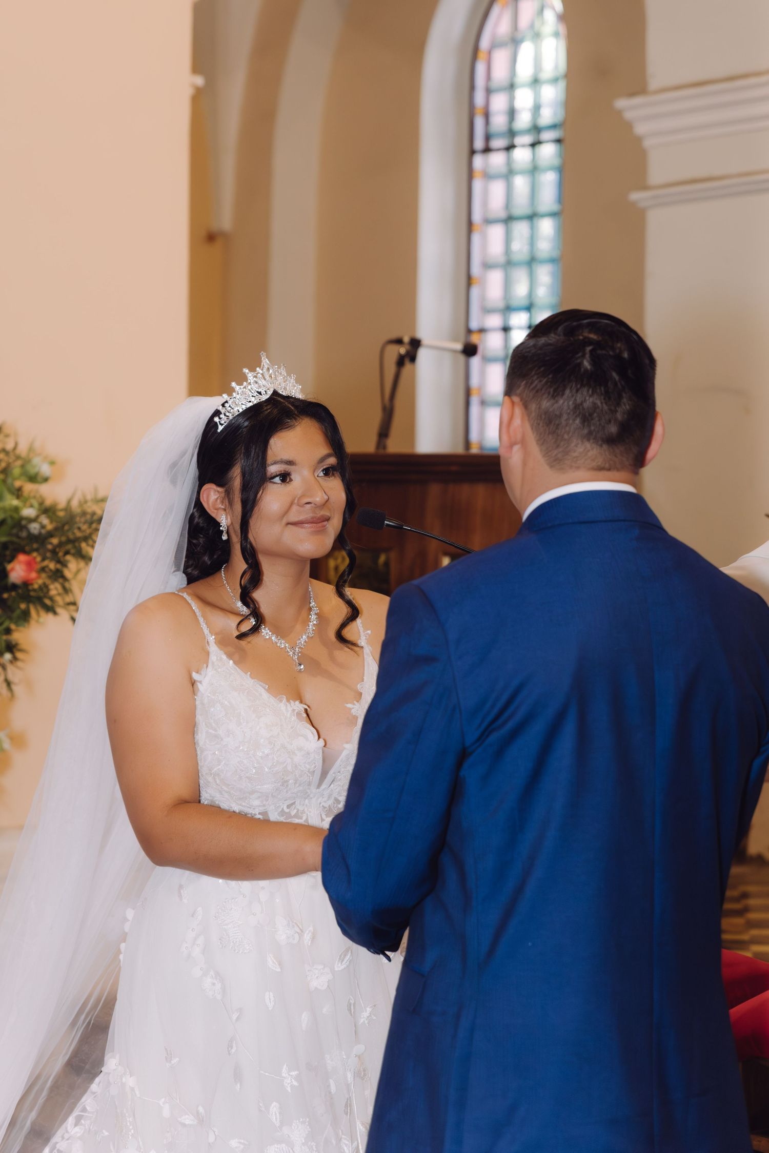 Bride in white lace gown and tiara smiles at groom in blue suit during church wedding ceremony.